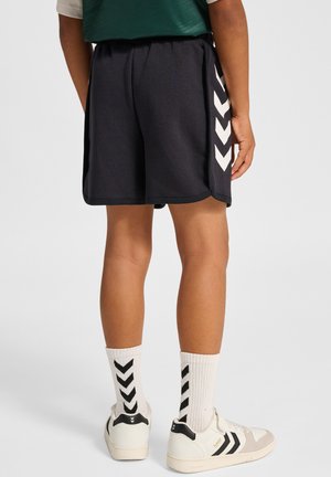 Marineblaue Shorts mit weißen Chevron-Streifen an der Seite, kombiniert mit weißen Socken mit schwarzen Chevron-Mustern und cremefarbenen Sneakern mit schwarzen Akzenten.