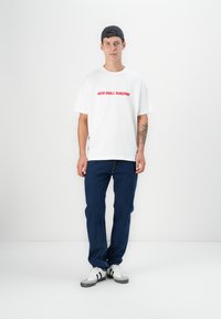 Hvitt kortermet t-skjorte med "RED BULL RACING" i rød skrift, kombinert med blå jeans og svart-hvite sneakers.