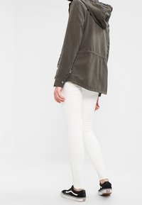 Veste à capuche vert foncé avec une texture lisse et des poches latérales, portée sur un jean skinny blanc et des baskets noires.