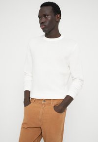 Νεαρός άνδρας με κοντά σγουρά μαλλιά που φοράει ένα άσπρο μακρυμάνικο ριμπέ σγουρό πουλόβερ και ανοιχτό καφέ παντελόνι, με τα χέρια στις τσέπες, κοιτάζοντας προς τα πλάγια.