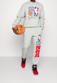 Sweat-shirt gris et pantalon de jogging avec des graphiques NBA 2022 en rouge et bleu, tenant un ballon de basket et portant des baskets rouges.