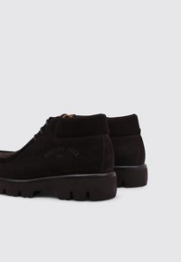 Botas de tobillo de ante marrón con superficie texturizada, que cuentan con una suela de goma negra gruesa y un logotipo en relieve en el talón.