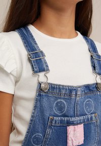 Pantalones de mezclilla con herrajes plateados, tirantes ajustables, parche rosa y patrones circulares. Combinados con una camiseta blanca de canalé.