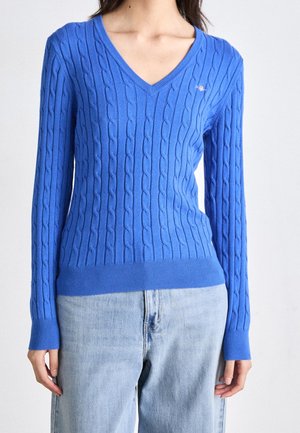 Kobieta ubrana w niebieski sweter warkoczowy z dekoltem w serek i jasne dżinsy, stojąca z rękami swobodnie opuszczonymi wzdłuż ciała.