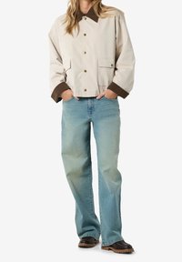 Person iført beige jakke med brun krave og manchetter, lyseblå lige jeans og sorte støvler med brune såler.