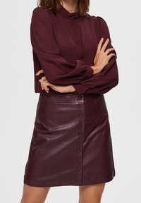 Blouse bourgogne avec des manches froncées et un détail plissé, associée à une jupe en cuir bourgogne ajustée avec des boutons sur le devant et une texture lisse.