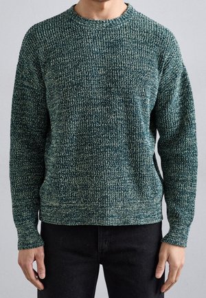 Uomo che indossa un maglione lavorato a maglia verde scuro e bianco e pantaloni neri, in piedi con le braccia rilassate lungo i fianchi.