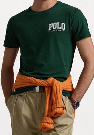 Mann trägt ein dunkelgrünes Polo Ralph Lauren T-Shirt, beige Hose, orangefarbenen Pullover um die Taille gebunden und eine braune Armbanduhr mit blauem Zifferblatt.