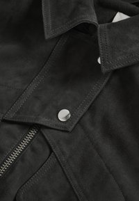 Primo piano del colletto di una giacca in camoscio nero con bottone a pressione argentato e dettaglio della cerniera metallica visibile.