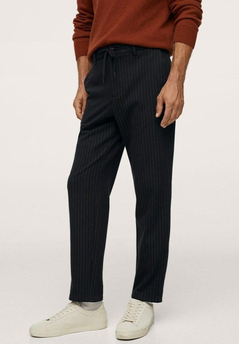 Homme portant un pantalon noir à rayures fines, un pull marron à manches longues, des chaussettes blanches et des baskets blanches, debout devant un fond uni.