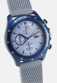 Reloj cronógrafo azul con una correa de silicona gris texturizada, que presenta una esfera azul claro, tres subesferas y un logo de Lacoste en la carátula.