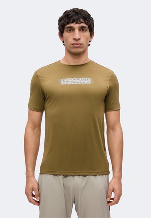 Hombre con camiseta de manga corta verde oliva con logo blanco de North Face y pantalones beige, de pie frente a un fondo claro y liso.