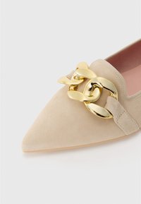 Pantof bej din suede, cu vârful ascuțit și un accent cu lanț aurit. Are un part superior moale și un detaliu cu curea texturată. Design simplu.