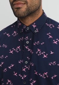 Marineblaues Hemd mit einem Button-Down-Kragen und einem rosa Vogel-Print. Der Stoff hat eine glatte Textur, graue Knöpfe und ein tailliertes Design.