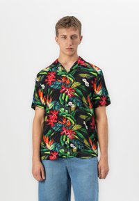 Jonge man met kort haar die een zwart overhemd met korte mouwen draagt, bedekt met rode en oranje tropische bloemmotieven, en lichte blauwe spijkerbroeken.
