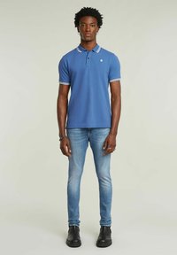 Blå polo med vit kant, kortärmad. Tillsammans med ljusa denimjeans och svarta ankelskor. Modellen står mot en neutral bakgrund.