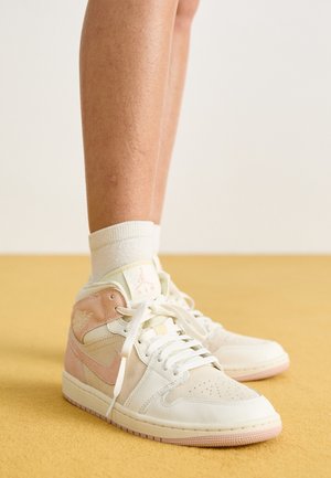 Osoba nosząca białe i jasnoróżowe buty Air Jordan z białymi skarpetkami, stojąca na żółtej podłodze na tle białej ściany.