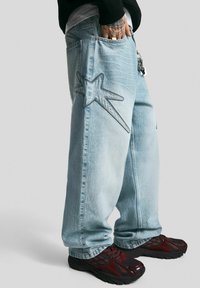 Light blue wide-leg jeans made of denim, featuring a prominent star stitched pattern on the back pocket. Black and burgundy athletic shoes.
