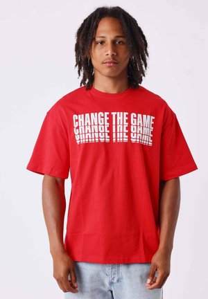 Jonge man met halflange dreadlocks, met een rood T-shirt met witte tekst "CHANGE THE GAME" en lichtblauwe spijkerbroek tegen een effen achtergrond.