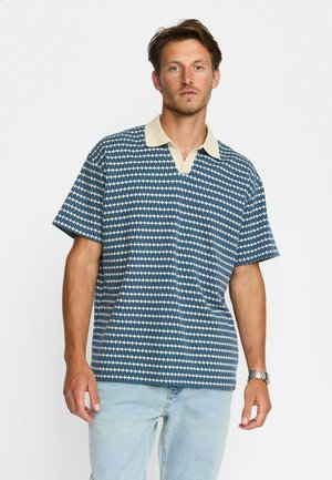 Man wearing a blue and white patterned short-sleeve polo shirt with a cream collar and light blue jeans, standing against a plain background.