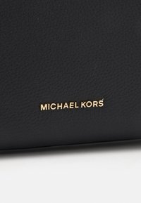 Sort pebret læder med en glat tekstur, prydet med et guldfolieret "MICHAEL KORS" logo.