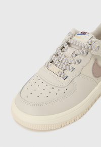Baskets Nike Force 1 beiges avec une boîte à orteils perforée, des lacets à motifs et une semelle de couleur claire. Dispose d'un logo discret et d'une tige en cuir lisse.