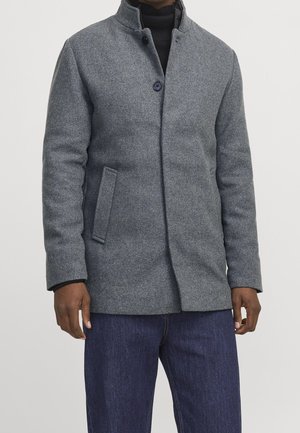 Homme portant un manteau en laine gris avec un col montant et une fermeture à boutons, associé à un jean bleu foncé et un pull col roulé noir.