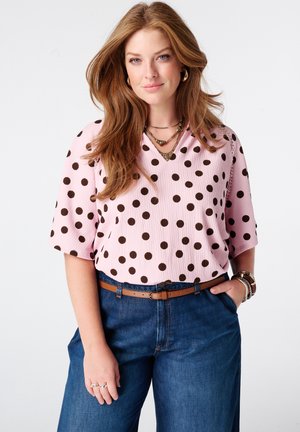 Femme aux longs cheveux roux portant un chemisier rose à manches courtes avec des pois marron, un jean bleu et des colliers superposés sur fond blanc.