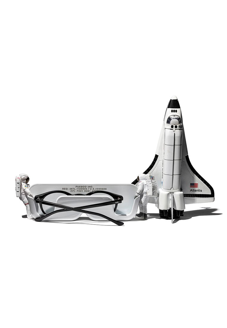 Weiße Brille mit rechteckigem Rahmen, schwarzen Akzenten und einem Modell des Space Shuttles mit der Bezeichnung "Atlantis" neben zwei Astronautenfiguren.