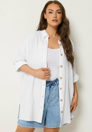 Vrouw met lang bruin haar draagt een witte blouse met knopen over een wit tanktop en lichtblauwe high-waist spijkershorts tegen een neutrale achtergrond.