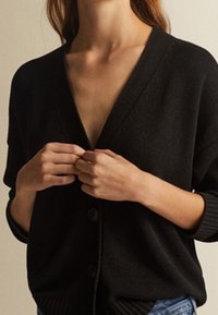 Schwarze Strickjacke mit V-Ausschnitt, langen Ärmeln und Knopfverschluss. Weicher Stoff mit gerippten Akzenten an den Ärmeln und am Saum.