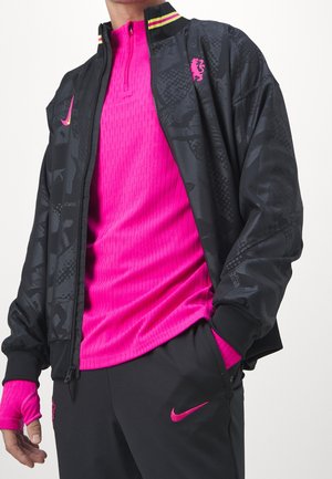 Personne portant une veste noire à motifs par-dessus un haut à fermeture éclair rose vif, un pantalon noir avec des logos roses et des gants roses sans doigts.