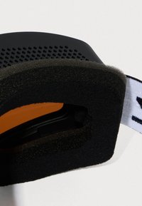 Czarne gogle sportowe z miękką, czarną piankową wyściółką wewnętrzną. Posiadają soczewki w pomarańczowym odcieniu widoczne wewnątrz oraz regulowany biały pasek.