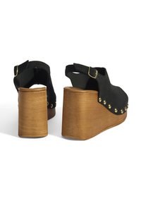 Schwarze Wildleder-Wedgesandaletten mit einem Holzabsatz, ausgestattet mit einem Schnallenriemen und goldenen Nieten entlang der Kante. Offenes Rückendesign für Atmungsaktivität.