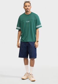 Grønn kortermet skjorte med hvite striper, kombinert med blå denimshorts. Brune joggesko og hvite sokker fullfører antrekket.