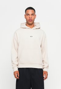 Jasno beżowa hoodie z kieszenią kangurzą, kapturem ze sznurkiem i subtelnym logo na środku. Miękki, fakturowany materiał z ryflowanymi ściągaczami.