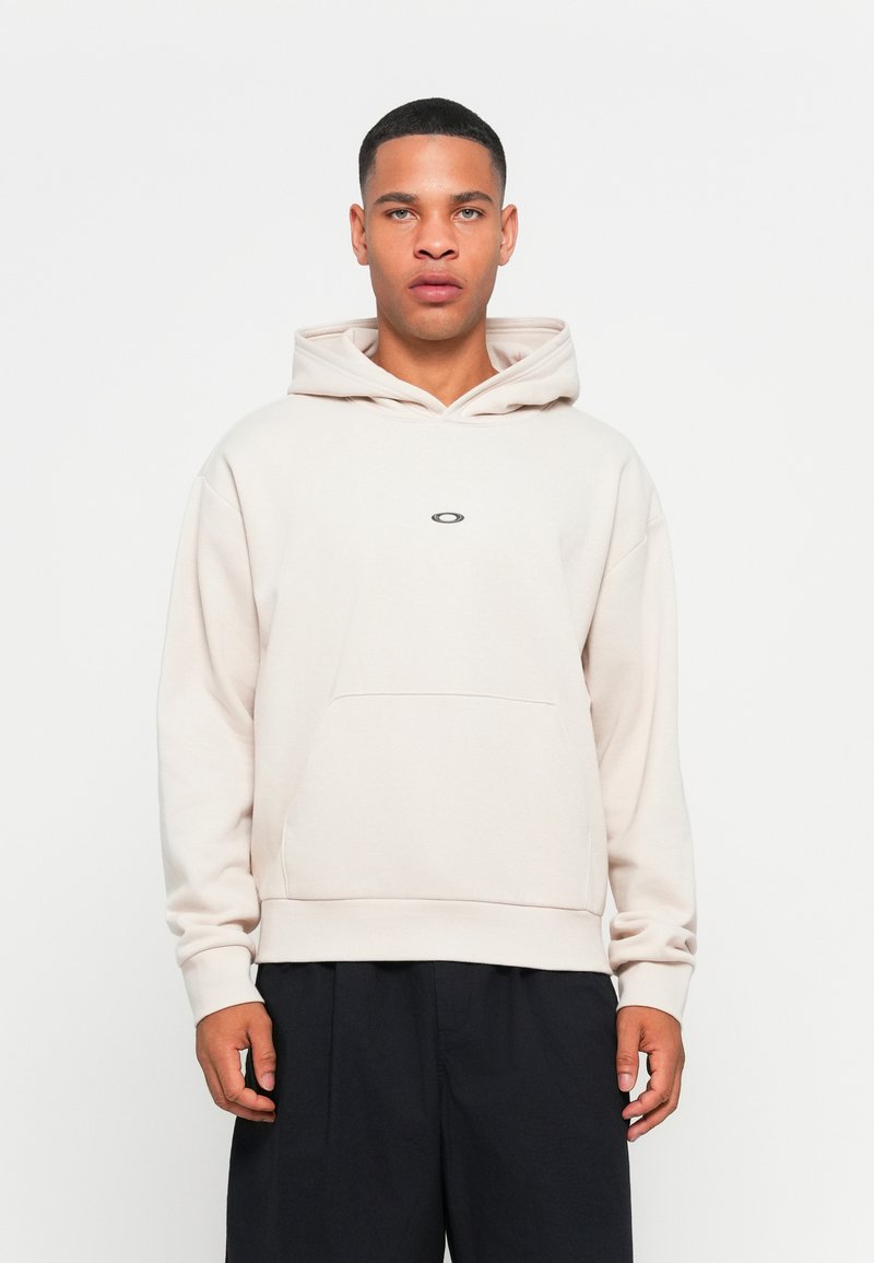 Jasno beżowa hoodie z kieszenią kangurzą, kapturem ze sznurkiem i subtelnym logo na środku. Miękki, fakturowany materiał z ryflowanymi ściągaczami.