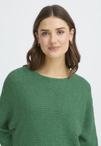 Pull en tricot vert avec une finition texturée et douce. Caractérisé par un large décolleté et une coupe décontractée. Design simple sans motifs ni accents supplémentaires.