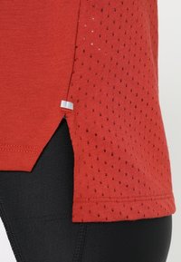 Punainen urheilupaita, jossa on hengittävä, rakennepintainen kangas, jossa on pieniä reikiä, sivuhalkioita ja tasainen helma. Mustat leggingsit näkyvät alla.