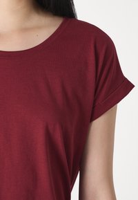 Burgundy színű pamut póló rövid, felgördített ujjakkal és kerek nyakkivágással, sima textúrával és a vállnál lazább szabással.