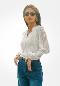 Donna che indossa occhiali da sole neri, un maglione lavorato a maglia bianco, jeans blu con grandi tasche e orecchini a forma di fiore in oro, in piedi con una mano sui fianchi.