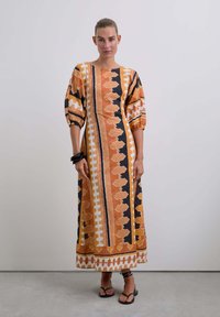 Robe maxi avec des motifs géométriques orange, noir et blanc. Manches bouffantes et coupe ample avec une silhouette droite. Des sandales complètent le look.