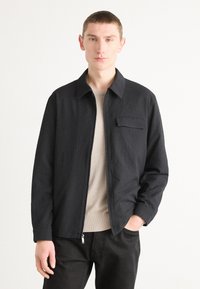 Black zip-up jacket with a textured fabric, pointed collar, and a single chest pocket, worn over a light beige sweater. Plain background.