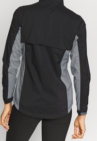 Svart och grå vattentät jacka med hög krage, långa ärmar och ett ventilerat bakpanel. Texturerad tyg genomgående.