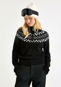 Sort sweater med hvid geometrisk design på bærestykket, halv-lynlås krave, ribbet nederkant, båret med sorte bukser og en cremet strikket hue.