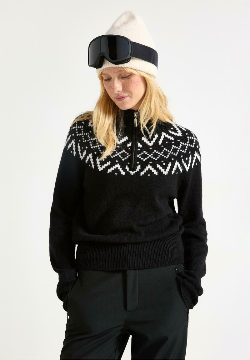 Sort sweater med hvid geometrisk design på bærestykket, halv-lynlås krave, ribbet nederkant, båret med sorte bukser og en cremet strikket hue.