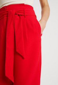 Pantalons larges rouges en tissu lisse, avec un détail de nœud à l'avant et des poches latérales. La taille est ajustée pour plus de structure.