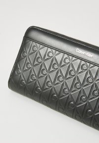 Black leather wallet featuring an embossed geometric pattern and a silver Calvin Klein logo. Zipper closure along the top edge.