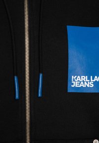 Crna zip-up hoodie s plavom logotipom sprijeda koji glasi "KARL LAGERFELD JEANS." Sadrži srebrni patentni zatvarač i plave vrhove pertli.