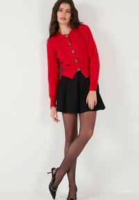 Roter gestrickter Cardigan mit tailliertem Schnitt und sechs dekorativen silbernen Knöpfen, kombiniert mit einem schwarzen schwingenden Rock und schwarzen High Heels.